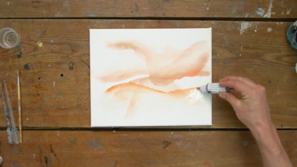 Vista superior de artista masculino pinta un cuadro abstracto, utiliza pintura naranja sobre lienzos húmedos y distribuirlo con pincel — Vídeo de stock