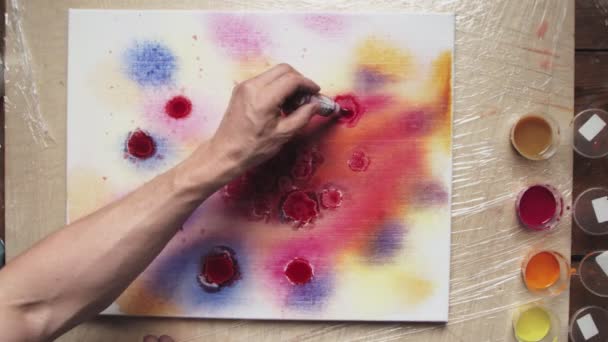 男性アーティストのトップビューは、パステルカラーの絞り染めやパステルの渦の絵を描きます。彼はたくさんの赤いペンキをぬれたキャンバスに注ぎ、赤い円を作る — ストック動画