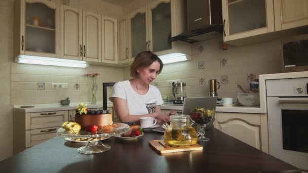 Kvinna dricker kaffe, äter tårta och typer på anteckningsbok i köket — Stockvideo