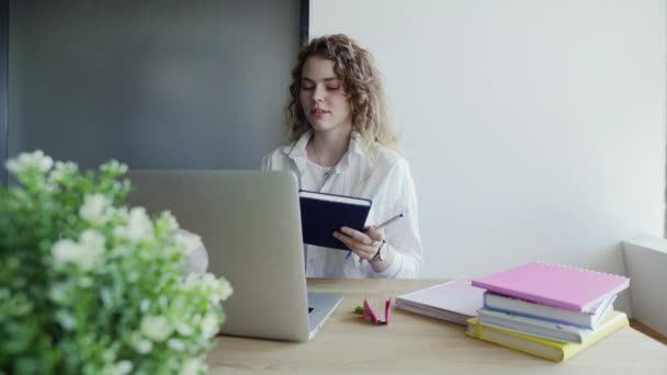 Mujer joven utiliza el ordenador portátil para charlas por chat de vídeo con sus colegas y toma notas — Vídeos de Stock