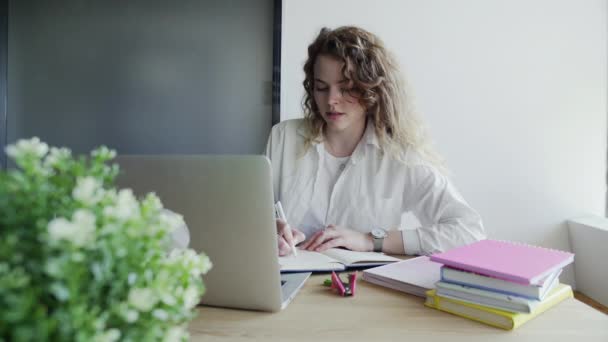 Young woman student uses laptop to works or study, she looks at screen and makes notes — Stock Video