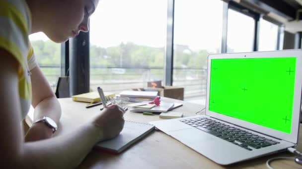 Вид сбоку на студентку, работающую на ноутбуке с зеленым экраном хромакея в домашнем офисе. Женщина смотрит на экран и делает заметки — стоковое видео