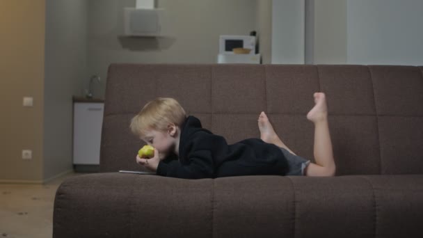 Lindo niño en negro sudadera con capucha se acuesta en sofá come una manzana y mira dibujos animados en la tableta — Vídeos de Stock