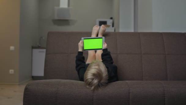 Lindo niño en sudadera con capucha negro pone en el sofá y utiliza la tableta con pantalla verde — Vídeo de stock
