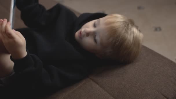 Κοντινό πλάνο του χαριτωμένου μικρού αγοριού με μαύρη κουκούλα ξαπλώνει στον καναπέ και χρησιμοποιεί tablet. Μια μικρή χειραψία — Αρχείο Βίντεο