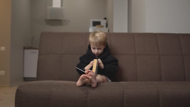 Menino bonito com capuz preto senta-se no sofá e lê grande livro interessante de contos de fadas — Vídeo de Stock