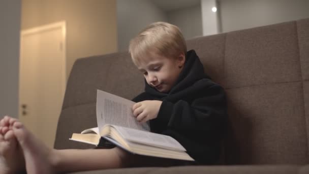 Menino bonito com capuz preto senta-se no sofá e lê livros. Menino vira páginas no sofá — Vídeo de Stock