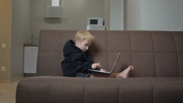 Menino bonito com capuz preto senta-se no sofá e usa notebook — Vídeo de Stock