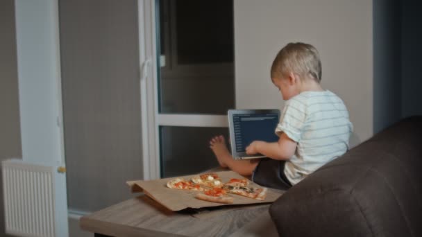 Vue arrière du jeune garçon assis sur la table avec ordinateur portable et mange de la pizza — Video