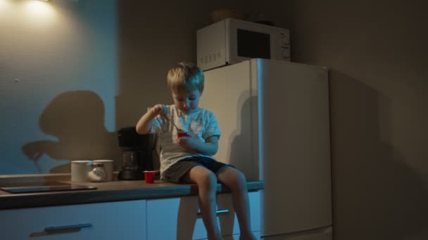 Mały chłopiec siedzi na stole w kuchni i je jogurt — Wideo stockowe
