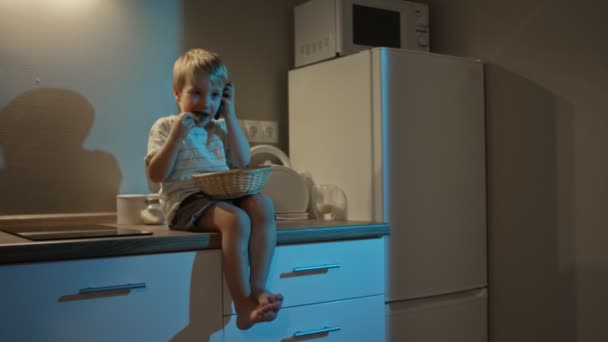 Niño feliz se sienta en la mesa de la cocina, come galletas y habla por teléfono por la noche — Vídeos de Stock