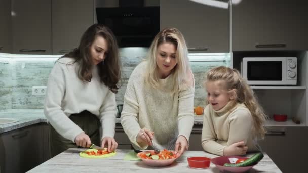 Νεαρή μητέρα και δύο κόρες της μαγειρεύουν σαλάτα στην κουζίνα. Η μητέρα αναμιγνύει σαλάτ ενώ η μεγαλύτερη κόρη της κόβει κόκκινη πιπεριά. — Αρχείο Βίντεο