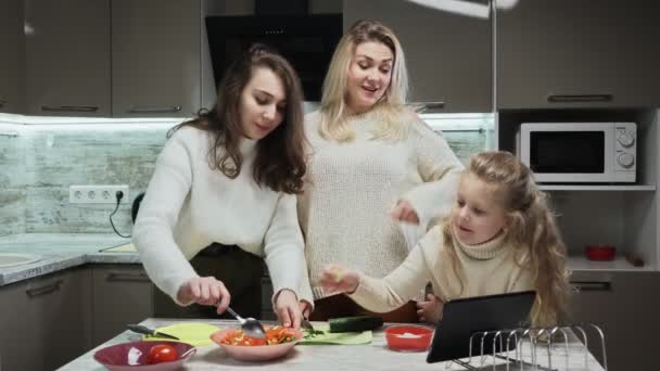 Νεαρή ευτυχισμένη οικογένεια, μητέρα και δύο κόρες μαγειρεύουν σαλάτα στην κουζίνα. Η μητέρα χρησιμοποιεί tablet με συνταγή νόστιμης σαλάτας με ντομάτες και αγγούρια — Αρχείο Βίντεο