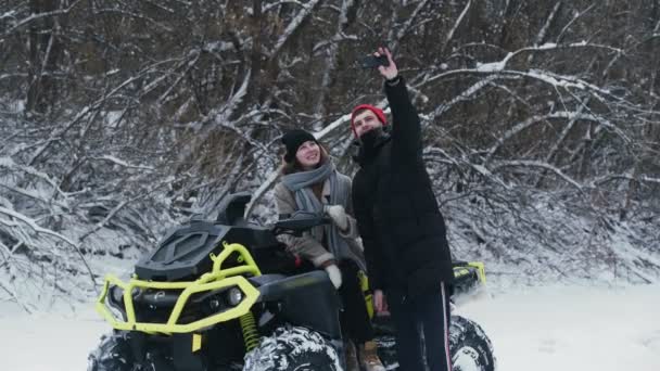 Fiatal pár pózol ATV Quad kerékpárral a téli erdőben. Egy piros kalapos fiatalember szelfit készít a barátnőjével az ATV-n. — Stock videók