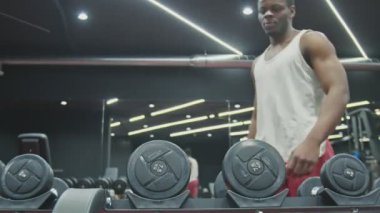 Genç afro adam spor salonunda iki dambılı alıp kaldırıyor.