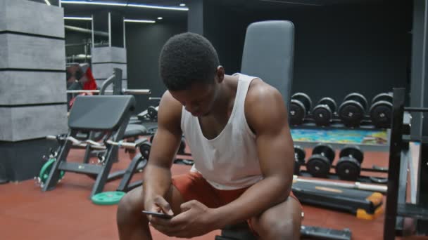 Pemuda berotot afro pria duduk di bangku, menggunakan telepon dan santai setelah latihan — Stok Video