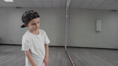 Beyaz tişörtlü ve siyah şapkalı küçük bir çocuk stüdyoda aynaya karşı hip hop yapıyor.