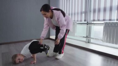 Beyaz tişörtlü küçük çocuk öğretmeninin yardımıyla yerde break dans yapıyor.