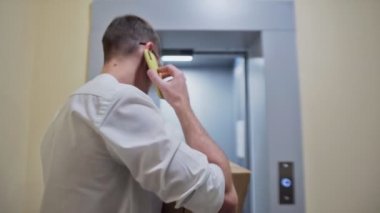 Gözlüklü genç kurye telefon kullanıyor, karton kutuyu tutuyor ve asansöre biniyor. Adam yeni bir daireye taşınıyor.