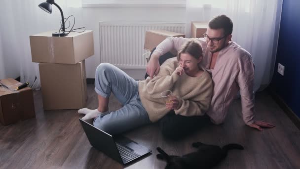 La pareja feliz se relaja después de mudarse a un nuevo hogar. Pareja se sienta en el suelo y ve la película de comedia en el portátil — Vídeo de stock