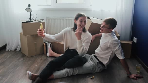 Glada par slappnar av efter att ha flyttat in i ett nytt hem. Par sitter på golvet och firar sin flytt i ett nytt hem. De spelar in video selfie på telefon — Stockvideo