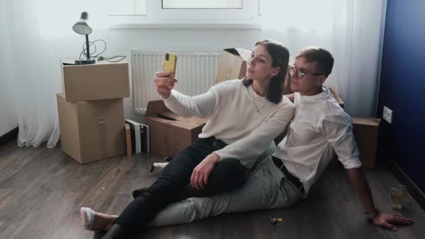 Glada par slappnar av efter att ha flyttat in i ett nytt hem. Par sitter på golvet och firar sin flytt i ett nytt hem. De tar en selfie på telefon — Stockvideo