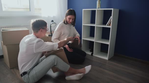 Счастливая пара открывает распакованные картонные коробки после переезда, семья сидит на полу в гостиной в день переезда в новый дом — стоковое видео