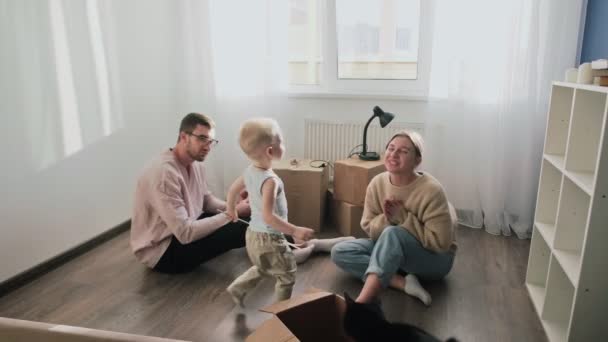 La pareja feliz se relaja después de mudarse a un nuevo hogar. Pareja sentada en el suelo, niño pequeño besa a su madre — Vídeos de Stock