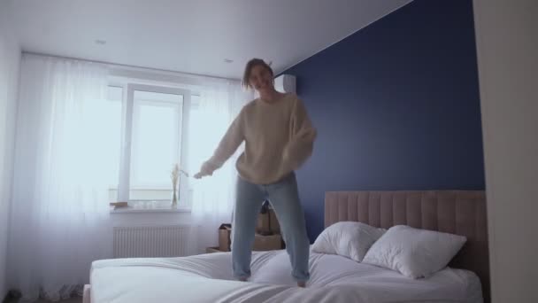 Happy young woman wearing jeans and sweater dances jumps on bed at home — Stock Video