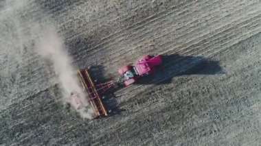 Kırmızılı bir traktörün havadan görünüşü, harrow sistemli tarla sürüşü, ekili tarlalar, arkasında toz izleri, yeni ekin ekimi için toprağı hazırlayan sütun.