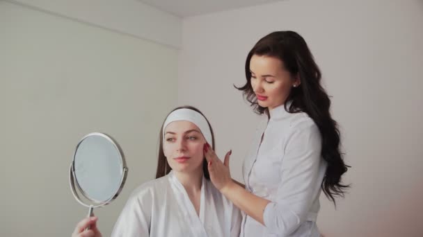 Cosmetoloog adviseert een vrouwelijke cliënt die naar spiegels kijkt in een schoonheidskliniek. Schoonheidssector concept. Schoonheidsspecialiste bereidt patiënt voor huidverzorging procedure in spa salon. — Stockvideo