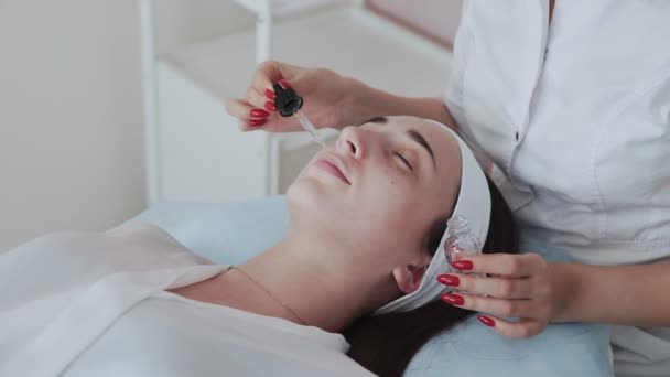 Kosmetolog nanáší peeling krém na ženskou klientskou tvář v kosmetickém salonu. Portrét samice dostane v lázních lékařskou kosmetickou proceduru na čištění obličeje. Kosmetický průmysl. Zpomalený pohyb — Stock video