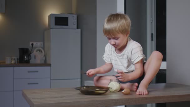 小さな男の子がテーブルの上に座り、砂糖を卵と一緒に皿に流し込み、混ぜ、味をつけます — ストック動画
