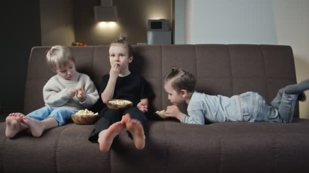 Tres hermanos se sientan en el sofá y comen palomitas en casa — Vídeo de stock