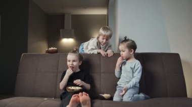 Üç kardeş kanepede oturup evde televizyon izlerken patlamış mısır yiyor.
