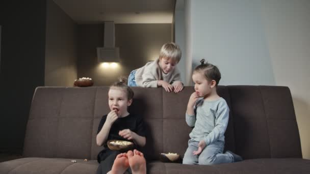 Trois frères s'assoient sur le canapé et mangent du pop-corn tout en regardant la télévision à la maison — Video