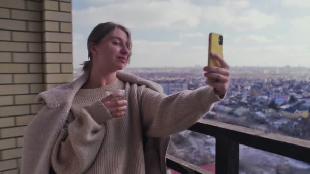 Ładna młoda kobieta na balkonie pije kawę lub herbatę, robi sobie selfie i rozmawia przez telefon — Wideo stockowe