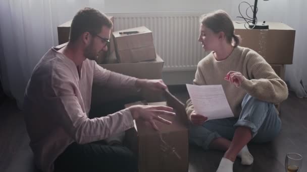 Casal feliz aberto desempacotando caixas de papelão após a realocação, família sentada no chão na sala de estar no dia em movimento em nova casa — Vídeo de Stock