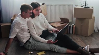 Mutlu çift yeni bir eve taşındıktan sonra rahatlar. Çift yerde oturuyor ve yeni evdeki yeni mobilyaları seçmek için dizüstü bilgisayar kullanıyor.