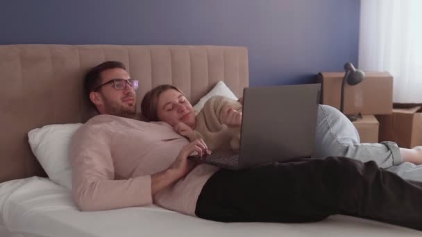 新しい家に引っ越した後、若いカップルはリラックスします。カップルはベッドで寝そべってノートブック画面を見る — ストック動画