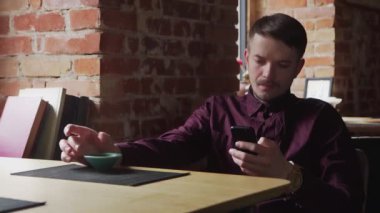Genç yakışıklı adam çay içiyor ve kafede cep telefonunu kullanıyor.
