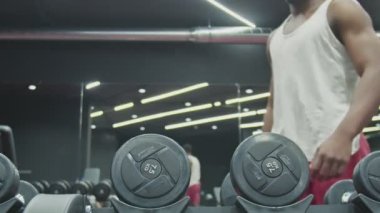 Genç afro adam spor salonunda iki dambılı alıp kaldırıyor.