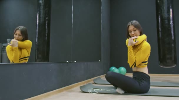 Молода жінка в жовтому верхньому сидить на килимку на підлозі і розтягує вправи — стокове відео