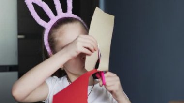 Pembe tavşan kulaklı küçük kız kırmızı kağıttan yapılmış makas nesneleriyle kesiyor. Mutfak masasında oturuyor..