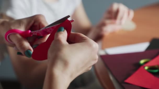 Close-up van de vrouw snijdt met schaar object eruit als lippen van een rood papier. Ze zit aan de keukentafel.. — Stockvideo