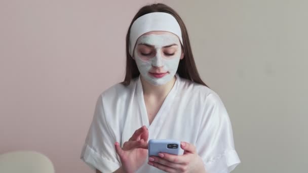 Wanita muda di salon kosmetologi kecantikan dengan masker tanah liat di wajah menggunakan telepon — Stok Video
