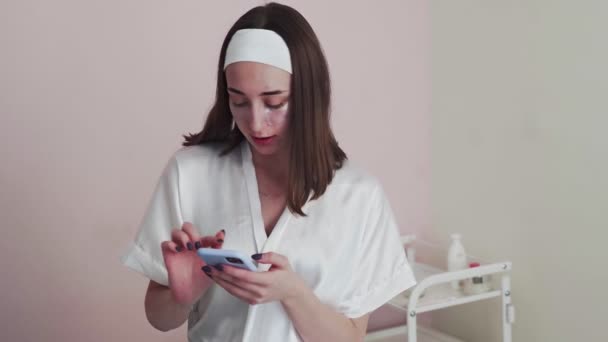 Молода жінка з колагеновими прокладками під очима використовує телефон у салоні краси . — стокове відео