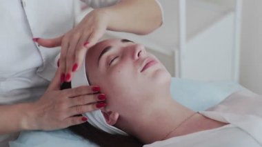 Güzellik uzmanının elleri kanepede gözleri kapalı yatan bir kadının yüzüne masaj yapıyor..