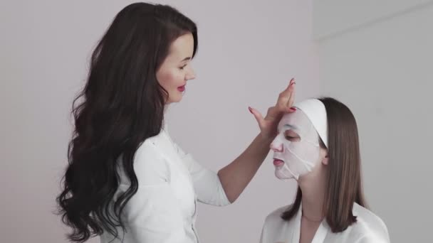 Wanita muda di salon kosmetologi kecantikan dengan topeng kain di wajah berbicara dengan dokter wanita dermatologis — Stok Video