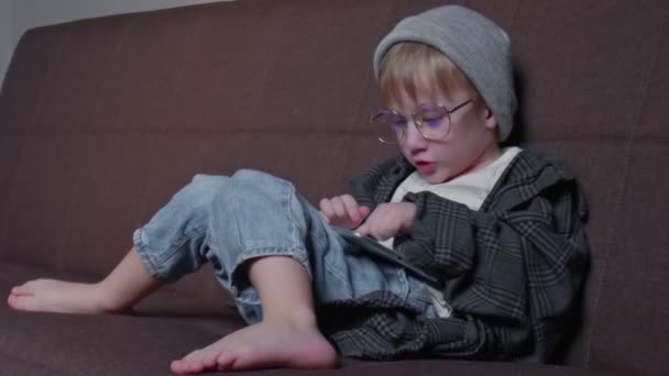 Hipster liten pojke Leker med surfplatta hemma. Kid Använda tablett sittande på en soffa — Stockvideo